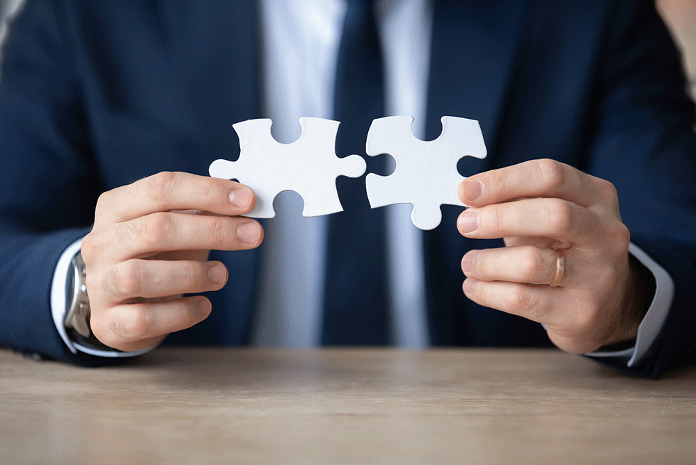 Detailný záber na podnikateľa skladajúceho dieliky skladačky – riešenie problému v biznise. Detailný záber na podnikateľa, ktorý drží v rukách dieliky puzzle a skladá ich, sedí pri pracovnom stole v kancelárii. Manažér alebo vedúci pracovník rieši problém, prijíma rozhodnutie – symbolika biznisového riešenia, spolupráce a spájania častí do jedného celku.